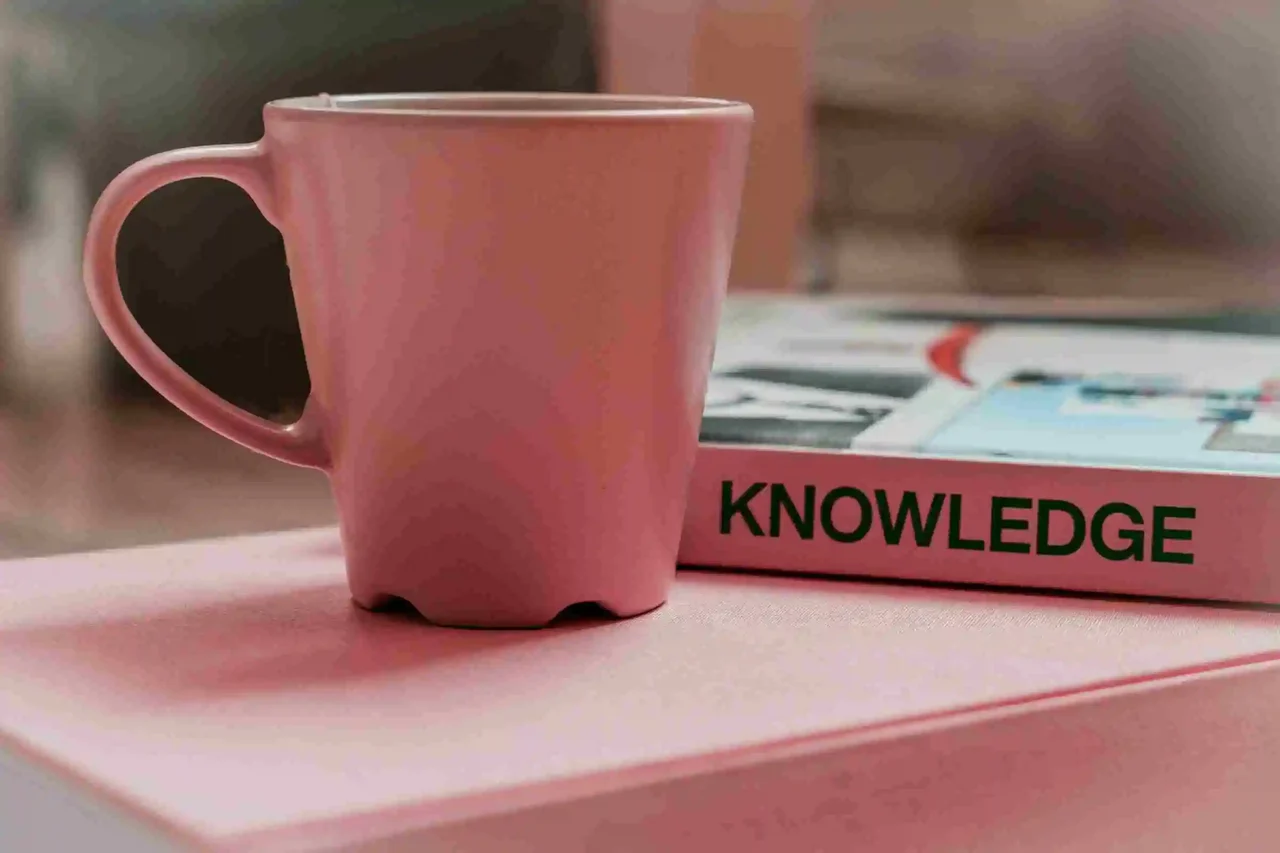 Pink ceramic mug next to a book titled “Knowledge” on a pastel surface.